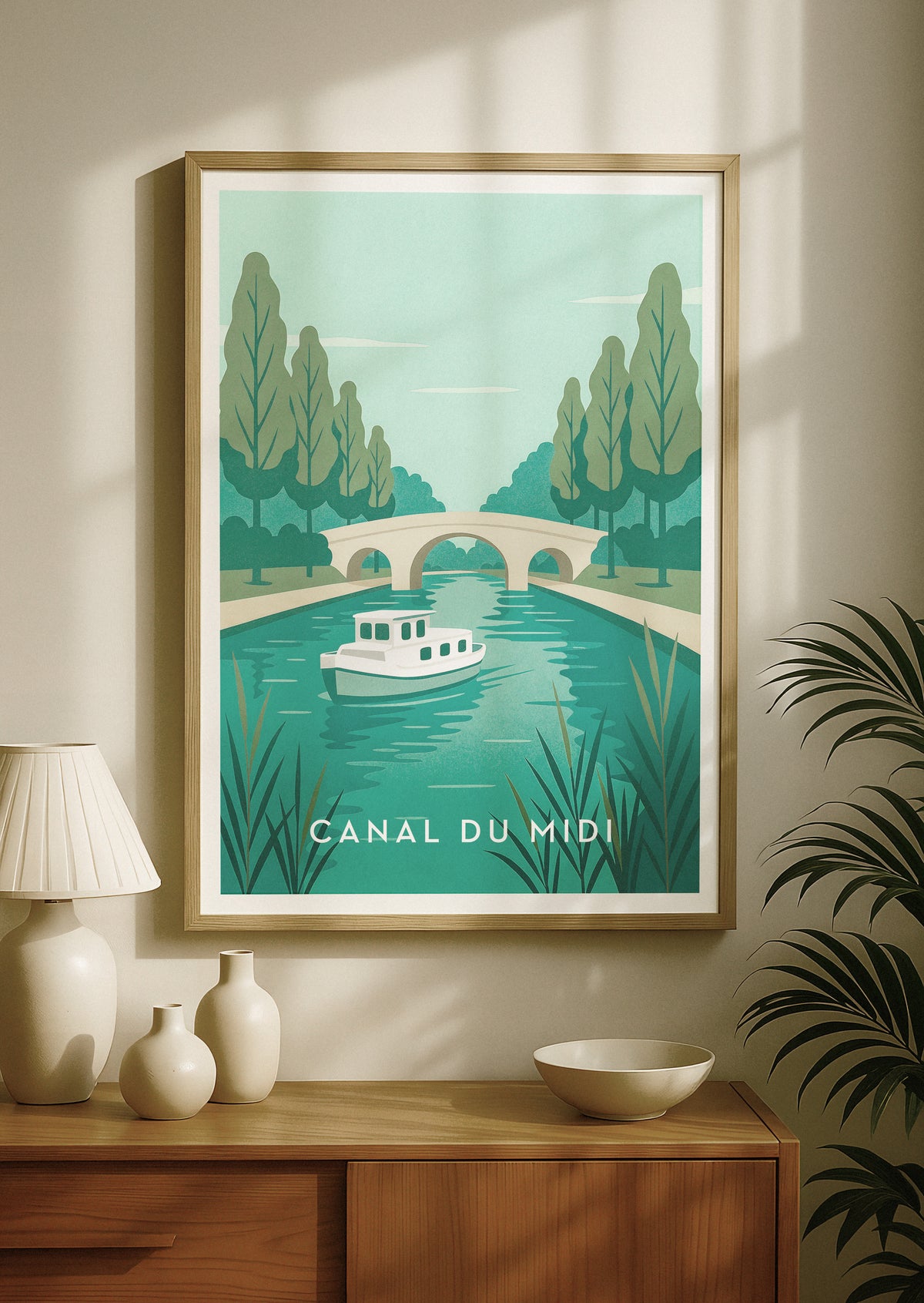 Affiche de voyage encadrée représentant le Canal du Midi en France, avec un bateau rétro sur une eau turquoise, des arbres et un pont de pierre, accrochée à un mur beige au-dessus d’une console en bois ornée de vases décoratifs et d’une lampe.